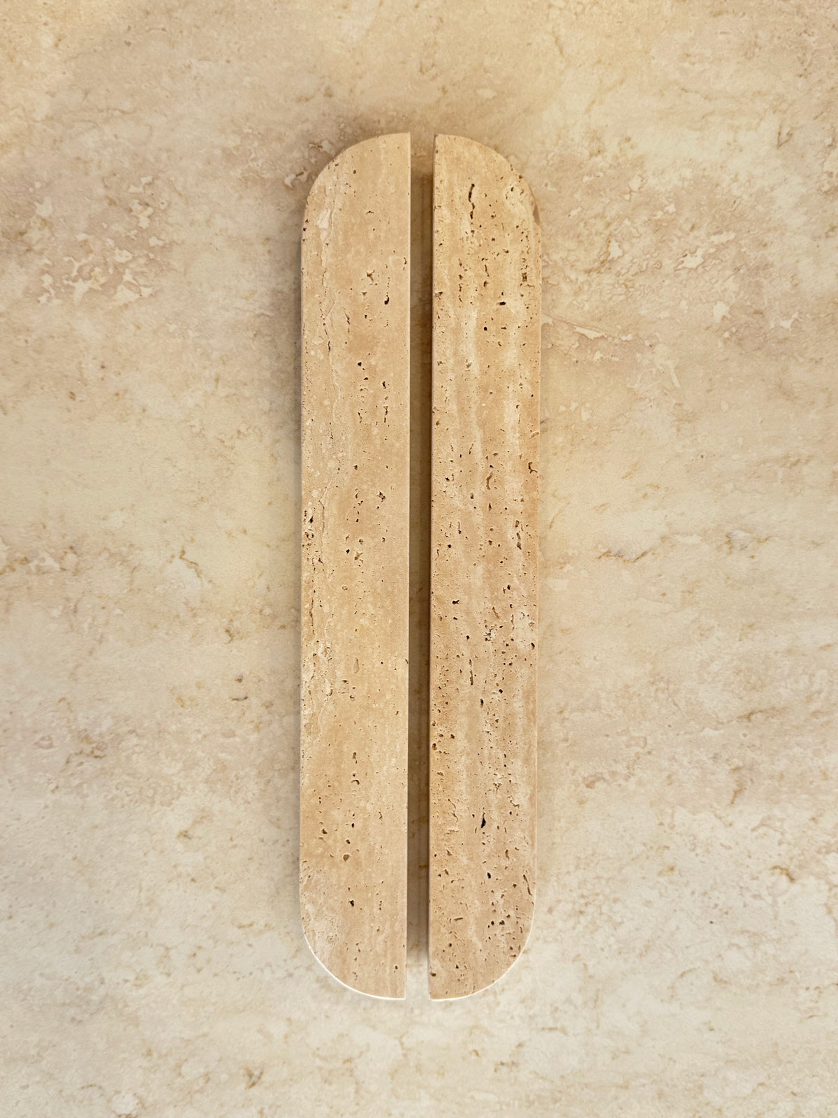 A minimalist, elongated oval wall-mounted light beige travertine stone sculpture with a central vertical fissure, set against a matching textured beige stone background.