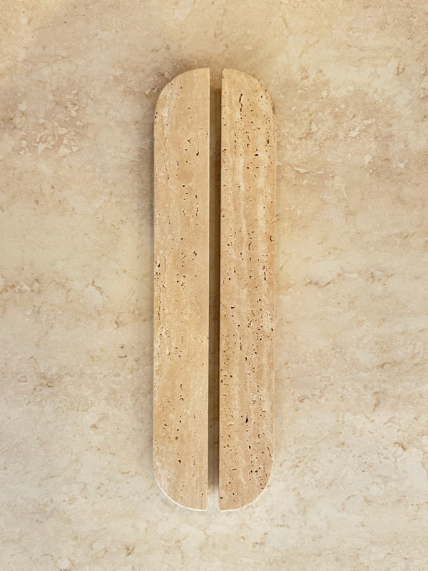 A minimalist, elongated oval wall-mounted light beige travertine stone sculpture with a central vertical fissure, set against a matching textured beige stone background.
