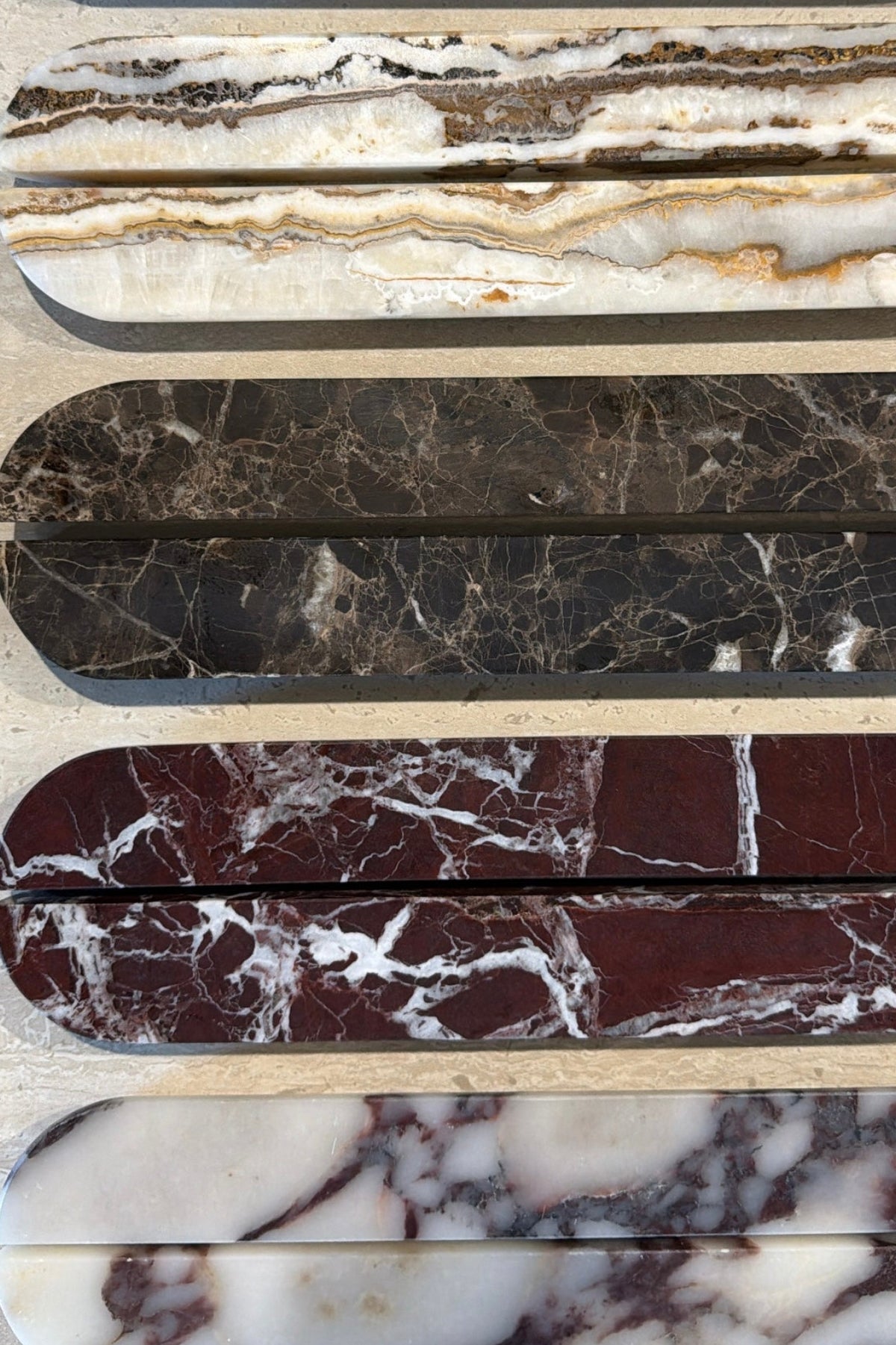 Six polished stone slabs with rounded ends are arranged vertically on a light surface. The stones show different marbled patterns and colors, including white and reddish-brown, cream and gold veining, dark gray and black marbling, deep burgundy and white veining, and white and gray marbling.
