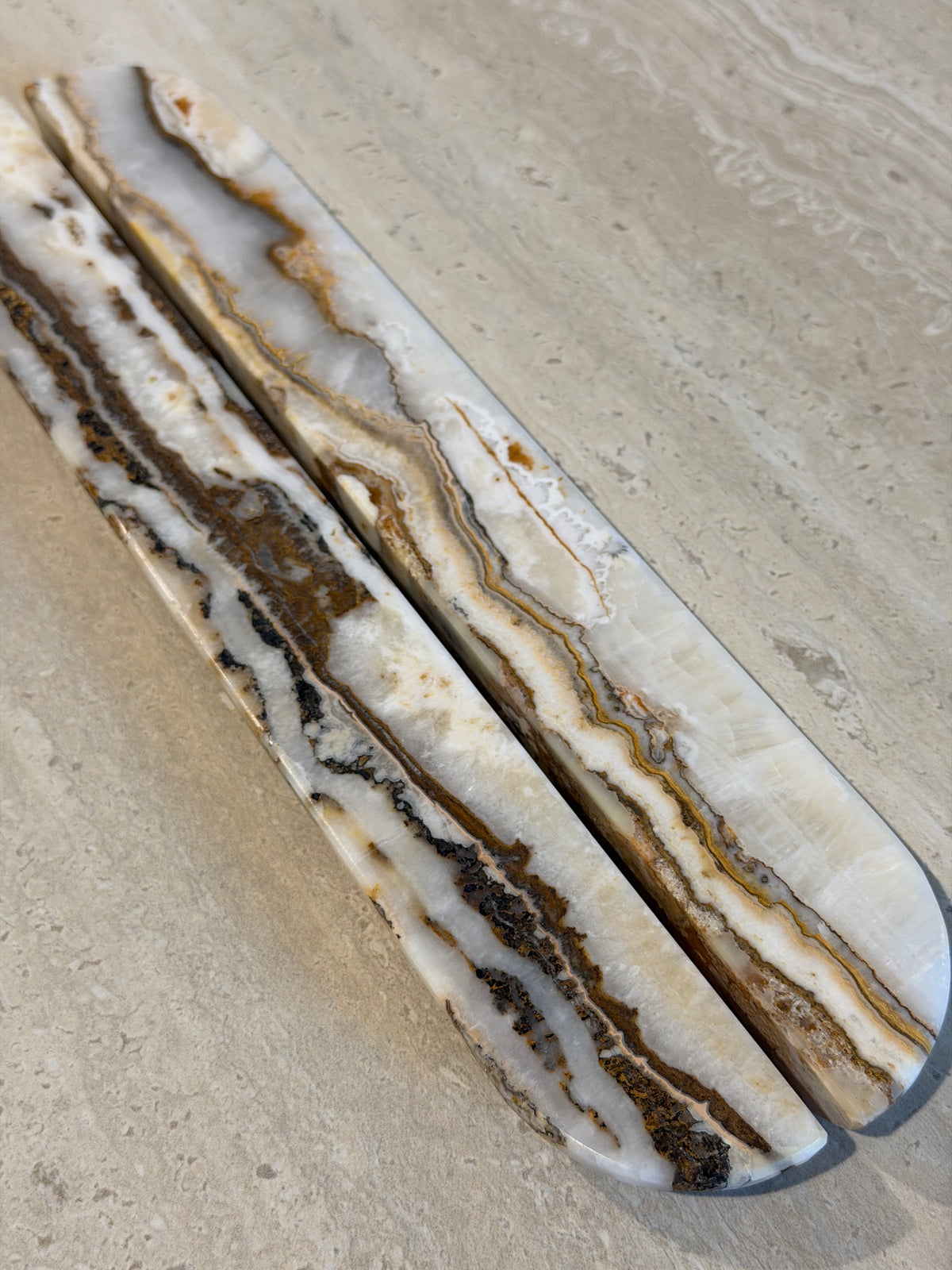Two elongated rounded bar pieces with natural agate-like banding in cream, white, tan, and dark brown, photographed on a light stone surface.