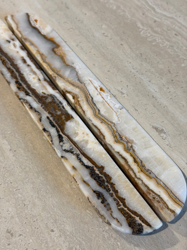 Two elongated rounded bar pieces with natural agate-like banding in cream, white, tan, and dark brown, photographed on a light stone surface.