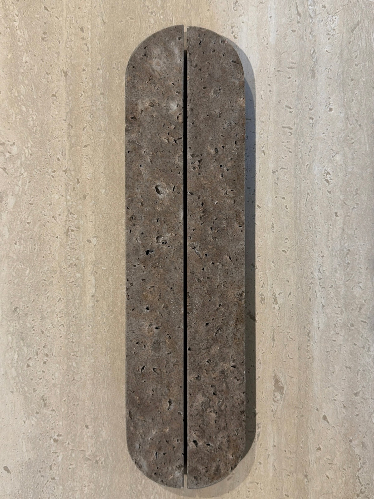 A long, narrow, rounded rectangular stone slab with a textured dark brown surface and a vertical center seam, mounted on a light beige stone wall.