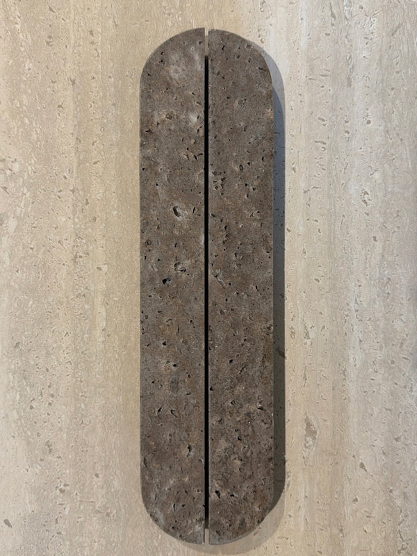 A long, narrow, rounded rectangular stone slab with a textured dark brown surface and a vertical center seam, mounted on a light beige stone wall.