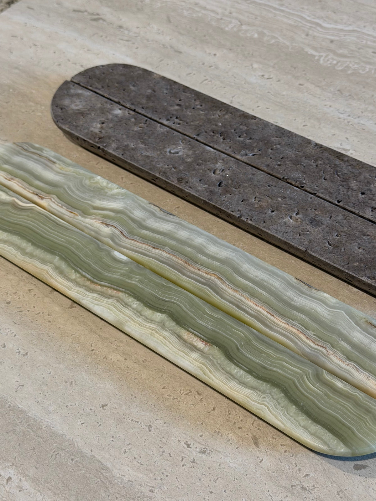 Two long rectangular stone slabs laid on a light beige stone surface. The slab in the foreground is pale green and cream with layered agate-like bands; the slab behind it is dark gray-brown with a rougher texture.