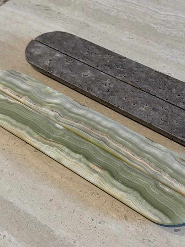 Two long rectangular stone slabs laid on a light beige stone surface. The slab in the foreground is pale green and cream with layered agate-like bands; the slab behind it is dark gray-brown with a rougher texture.