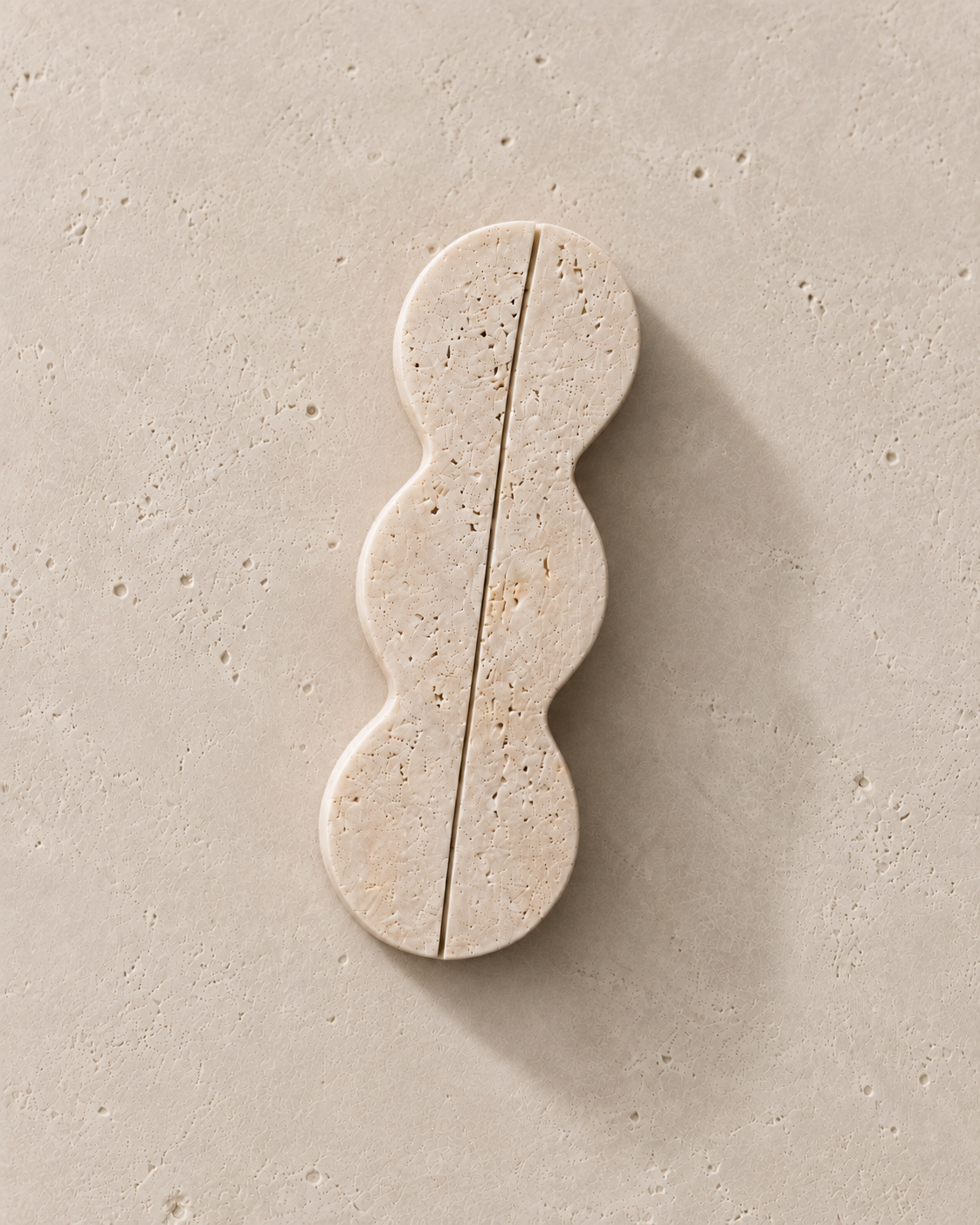 A single wavy, elongated stone tile or decorative slab in a warm beige travertine-like finish, photographed from above on a matching textured beige surface with a soft shadow.