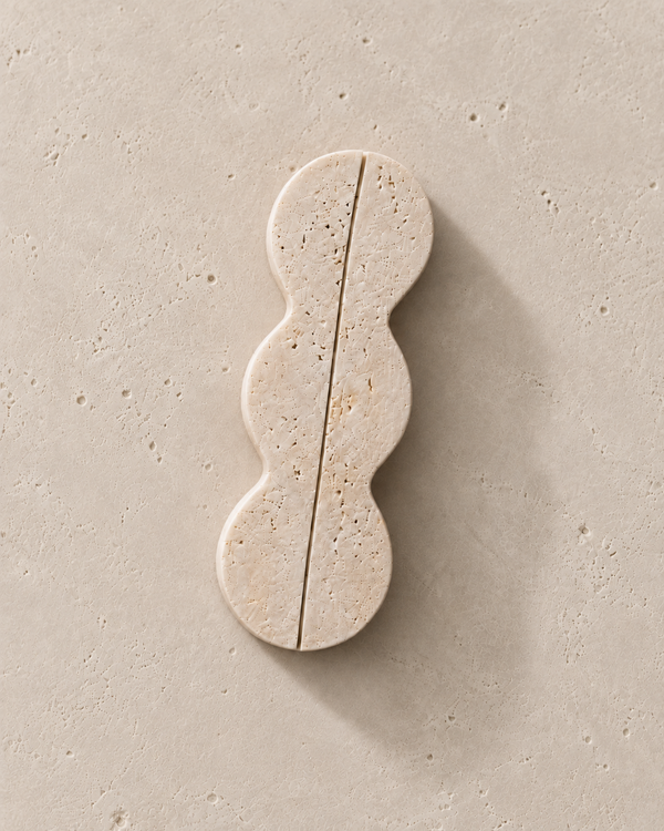 A single wavy, elongated stone tile or decorative slab in a warm beige travertine-like finish, photographed from above on a matching textured beige surface with a soft shadow.