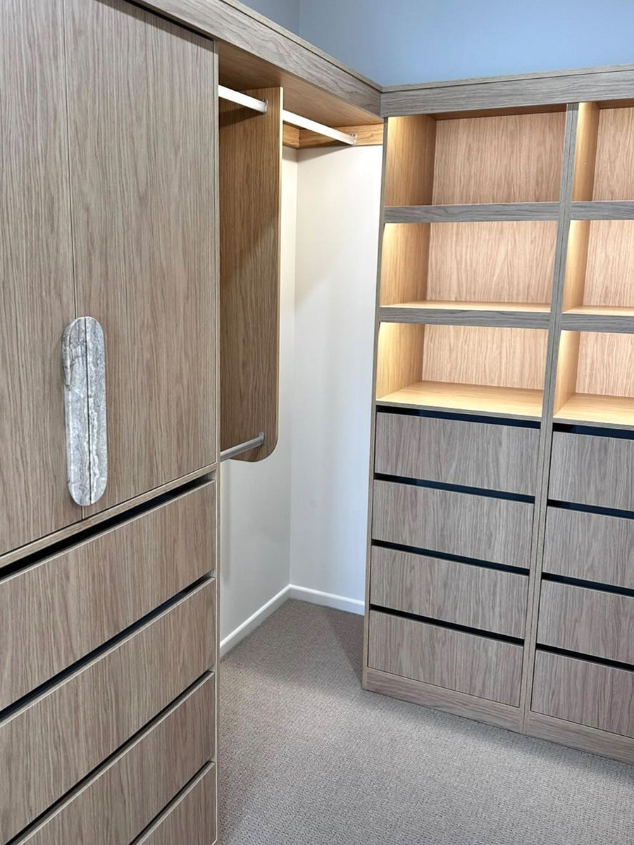Custom walk-in closet cabinetry with light oak wood grain finish, featuring drawers, open shelving with warm LED lighting, and a hanging rod area.