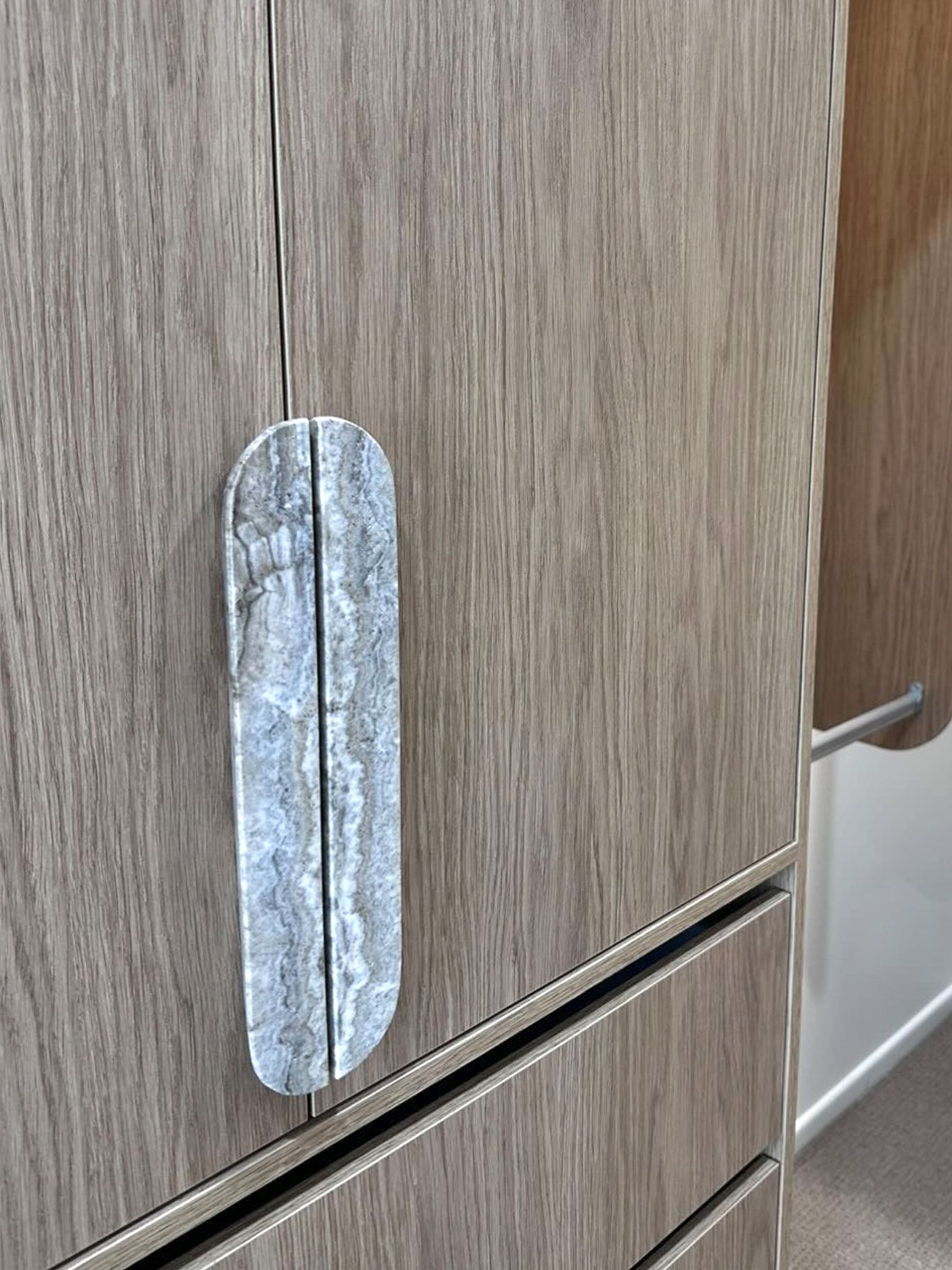 A close-up of a modern wardrobe or cabinet door with a light brown wood grain finish and a distinctive vertical oval handle made of polished gray marble.