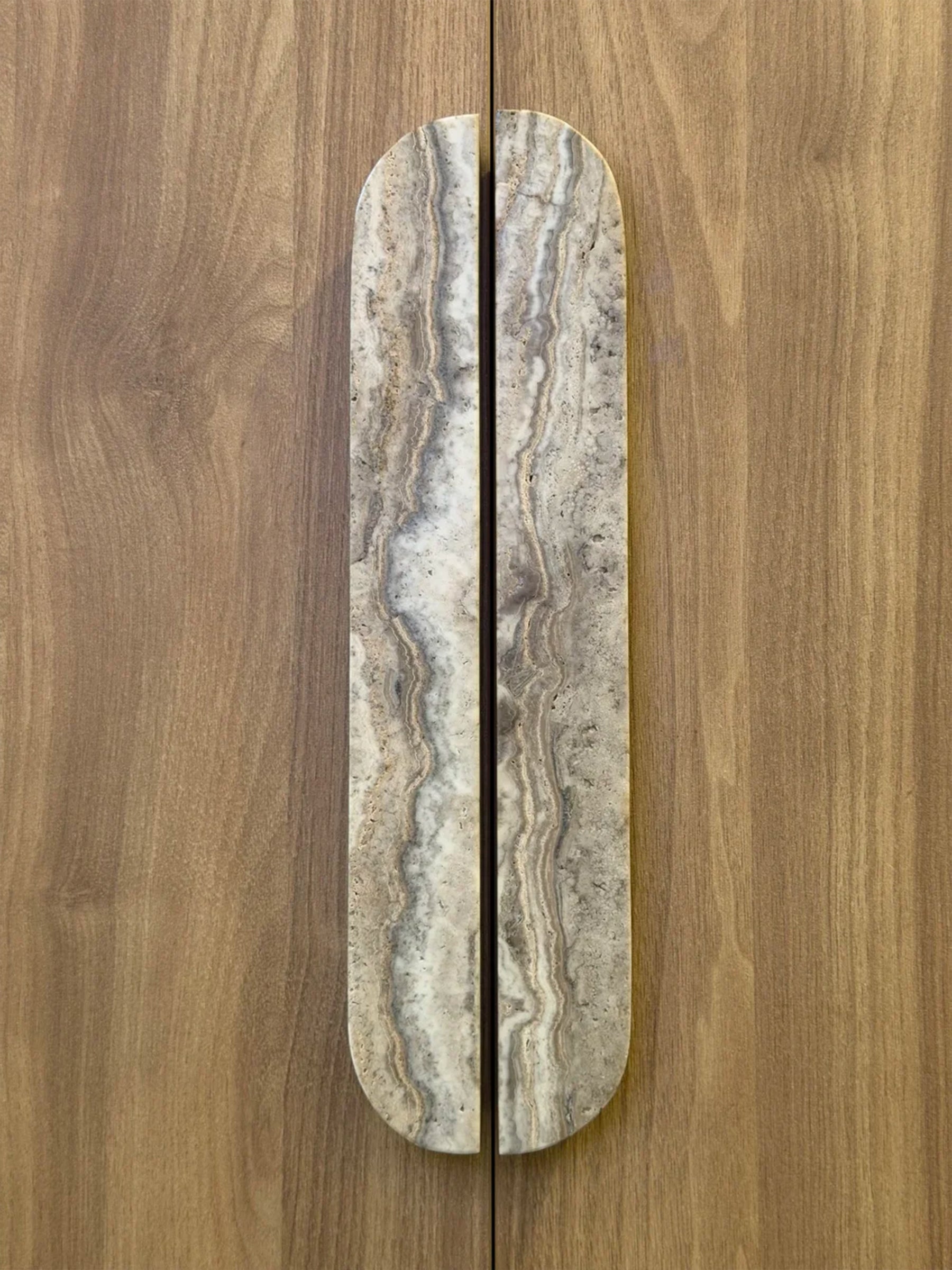 A long, narrow cabinet or door handle with a rounded top, made from polished stone with natural beige, gray, and brown veining, shown installed vertically on a wood-grain door.