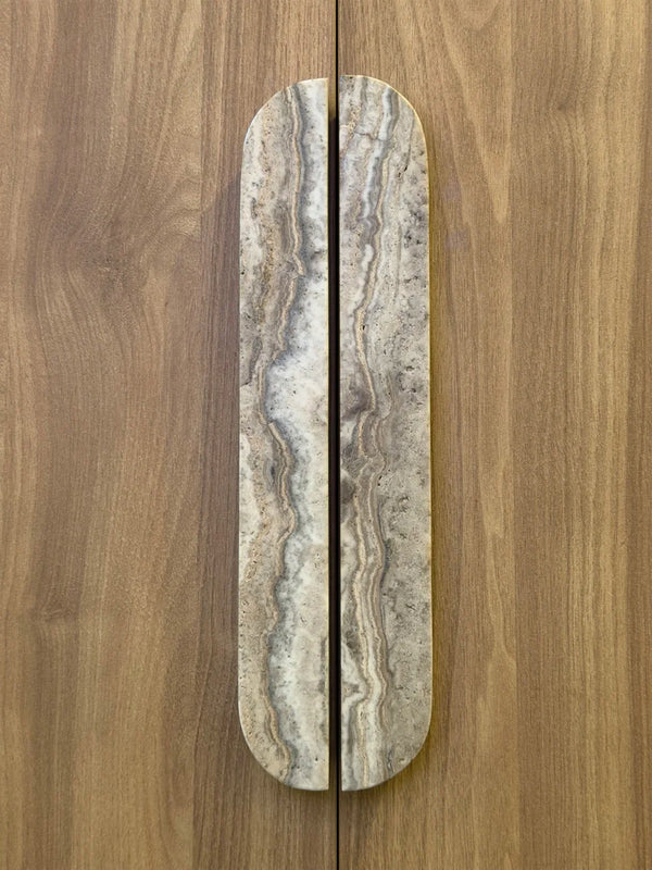 A long, narrow cabinet or door handle with a rounded top, made from polished stone with natural beige, gray, and brown veining, shown installed vertically on a wood-grain door.