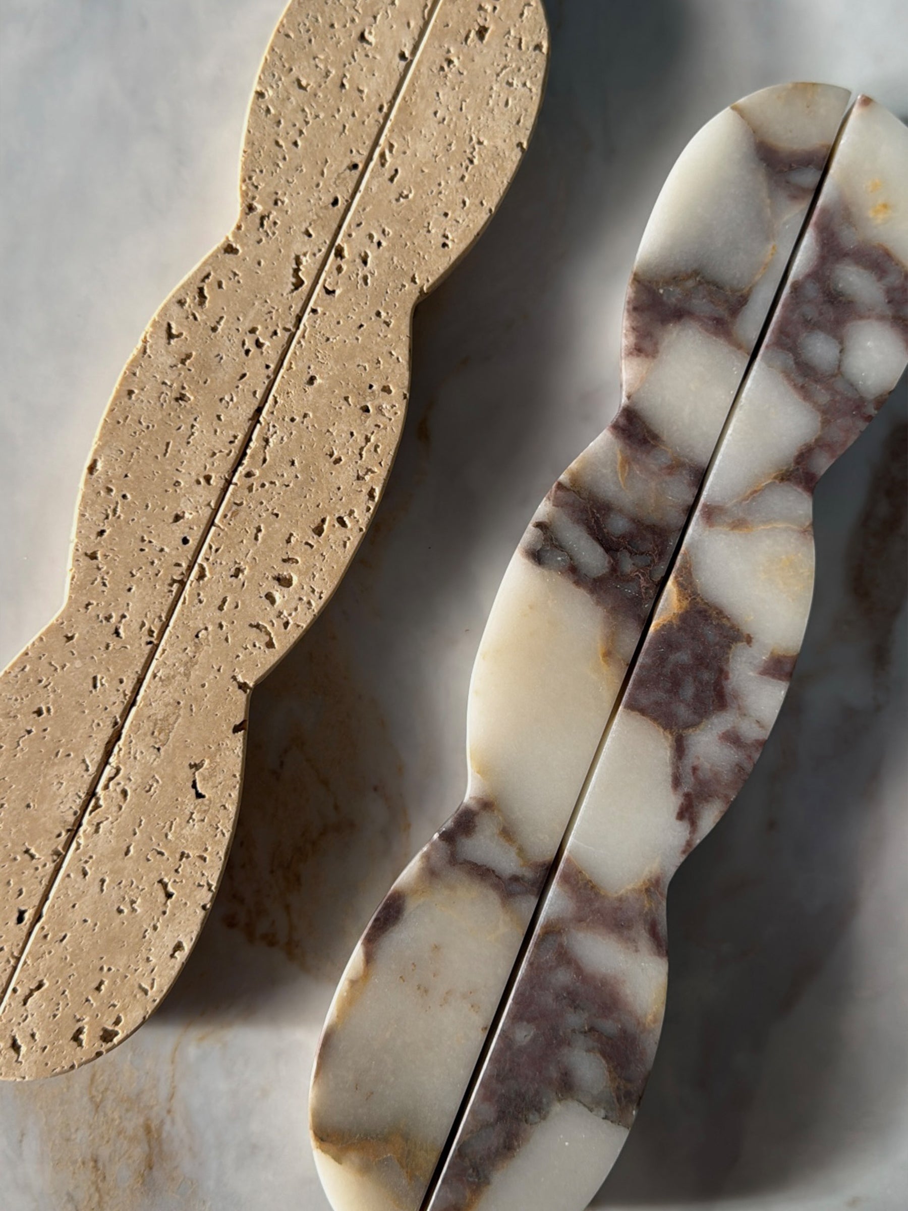 Two decorative soap bars with scalloped edges, one made of natural beige travertine and the other of white and brown marbled stone, displayed on a light marble surface.