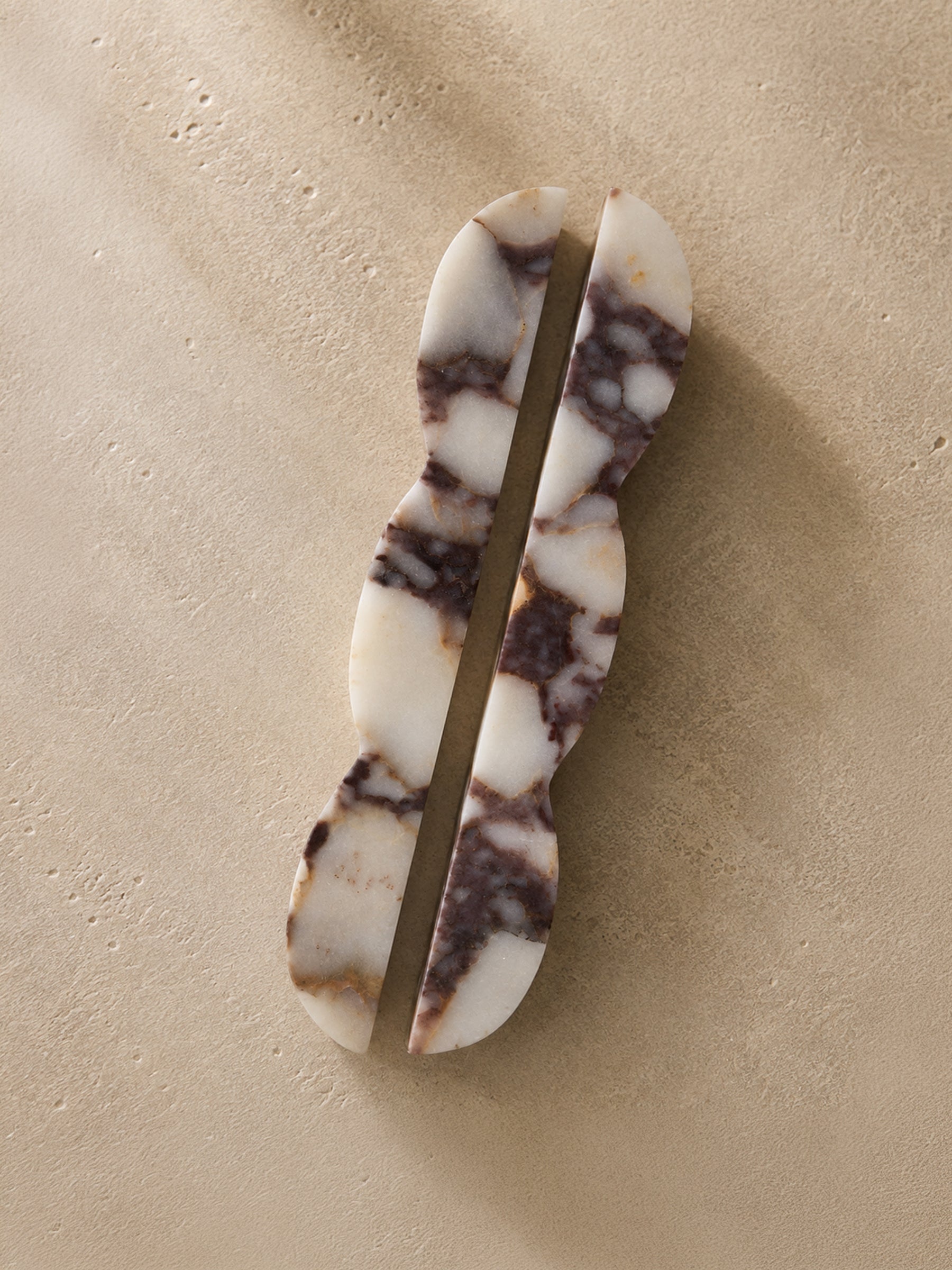 A pair of elongated, wavy hair clips made from white and dark brown marbled stone, displayed on a textured beige background.
