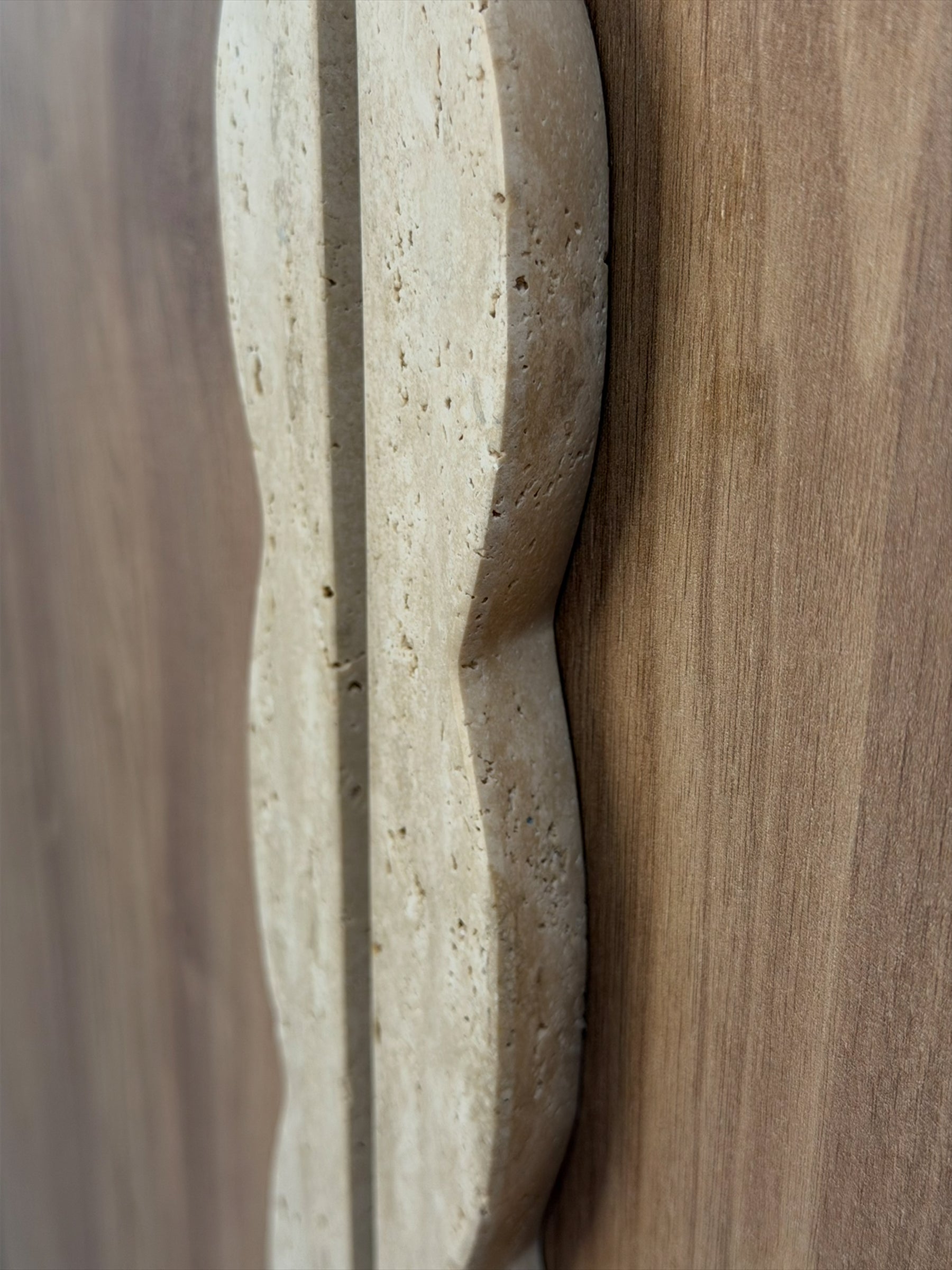 A close-up detail of a light beige travertine stone cabinet knob with a wavy, scalloped edge, set against a warm brown wood grain background.