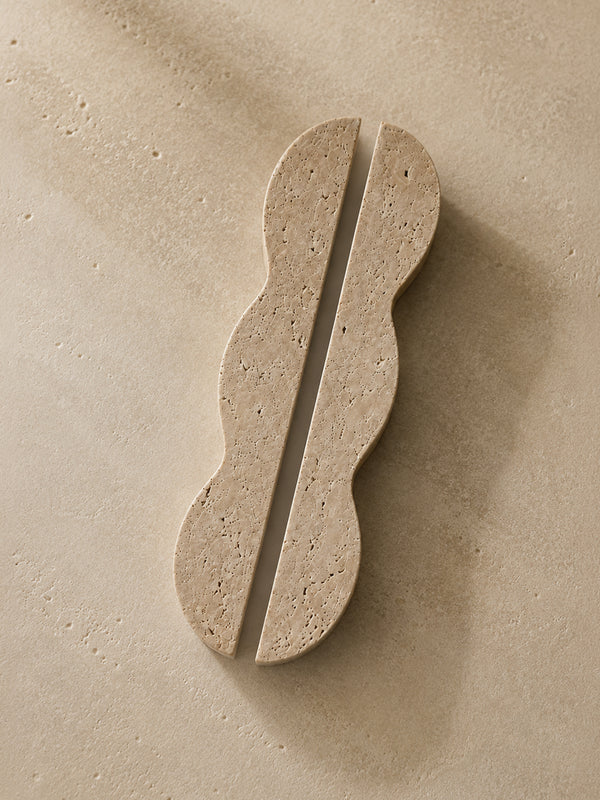 A pair of wavy-edged stone coasters in a light beige travertine-like material, displayed on a matching textured beige surface.