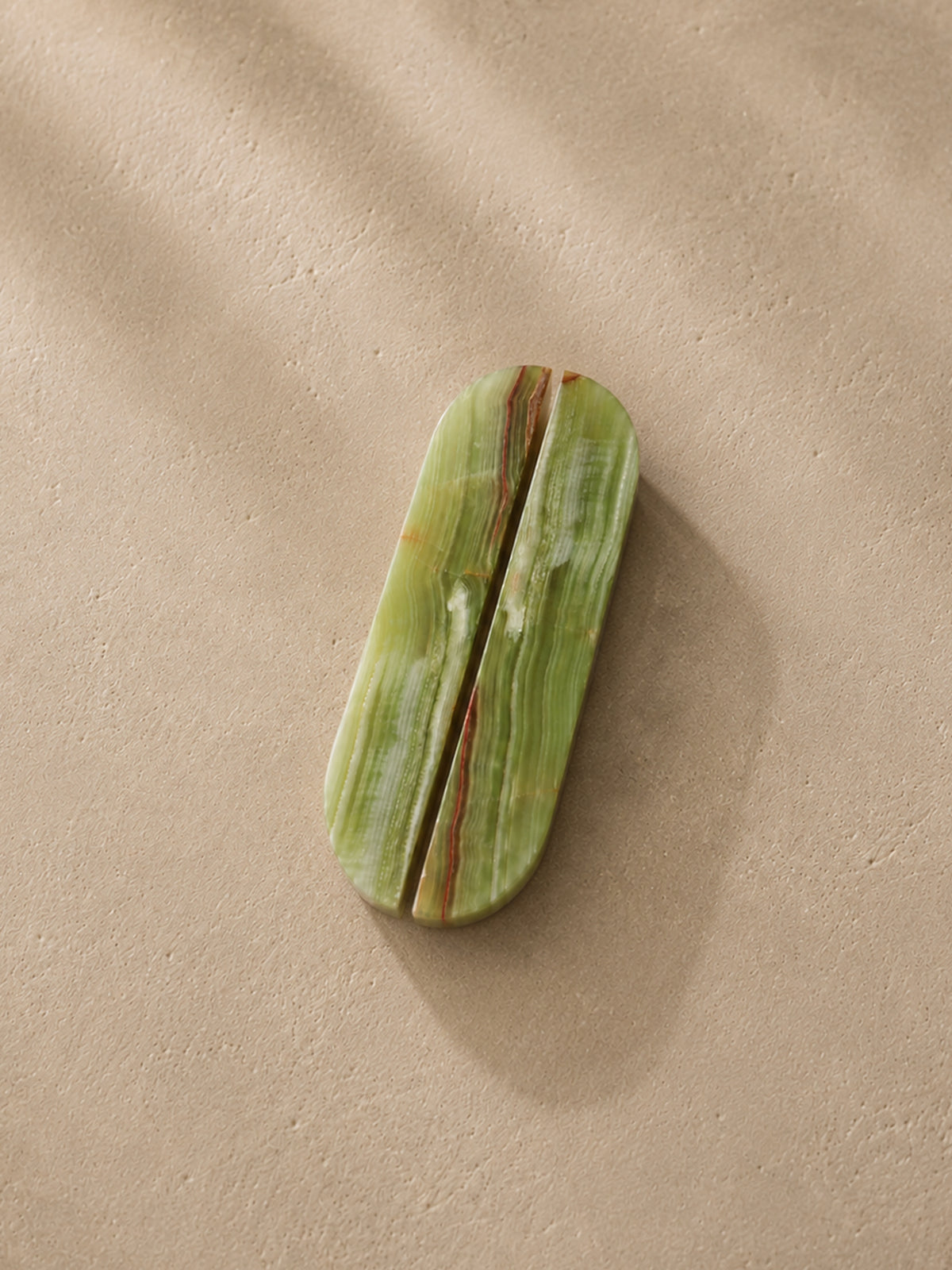 A polished green stone cosmetic case with a central split seam and natural marbled veining, resting on a textured beige surface with soft shadows.