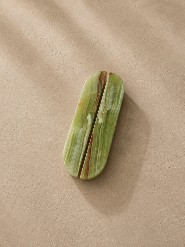 A polished green stone cosmetic case with a central split seam and natural marbled veining, resting on a textured beige surface with soft shadows.