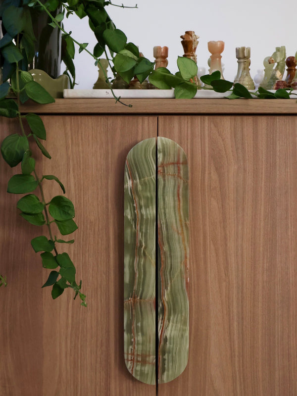 A decorative cabinet door handle made of green agate with natural banding and subtle orange-red veining, mounted on a wooden cabinet with a green trailing plant and chess pieces above.