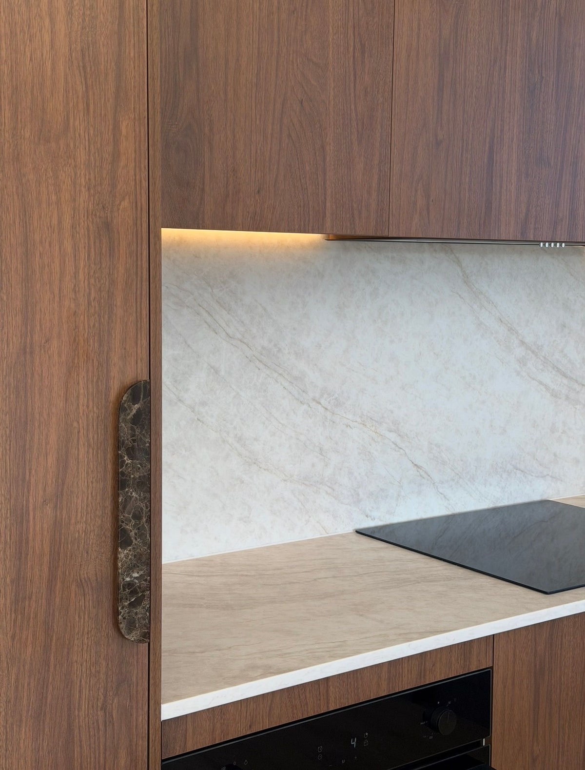 Close-up of a modern kitchen cabinet with a dark wood grain finish, a vertical black marble handle, a light beige marble backsplash, and a black built-in cooktop.