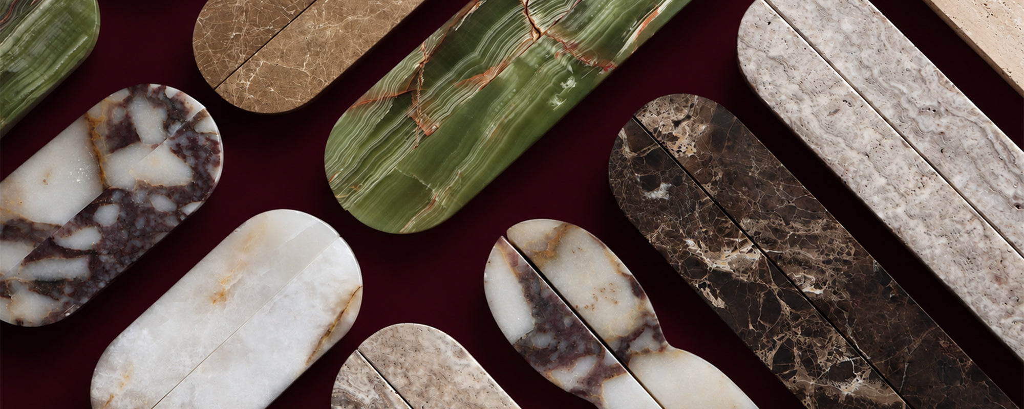 A close-up flat lay of multiple polished stone slabs or tiles in varied colors and marbled patterns, including green, cream, brown, black, and gray, arranged diagonally on a dark burgundy background.