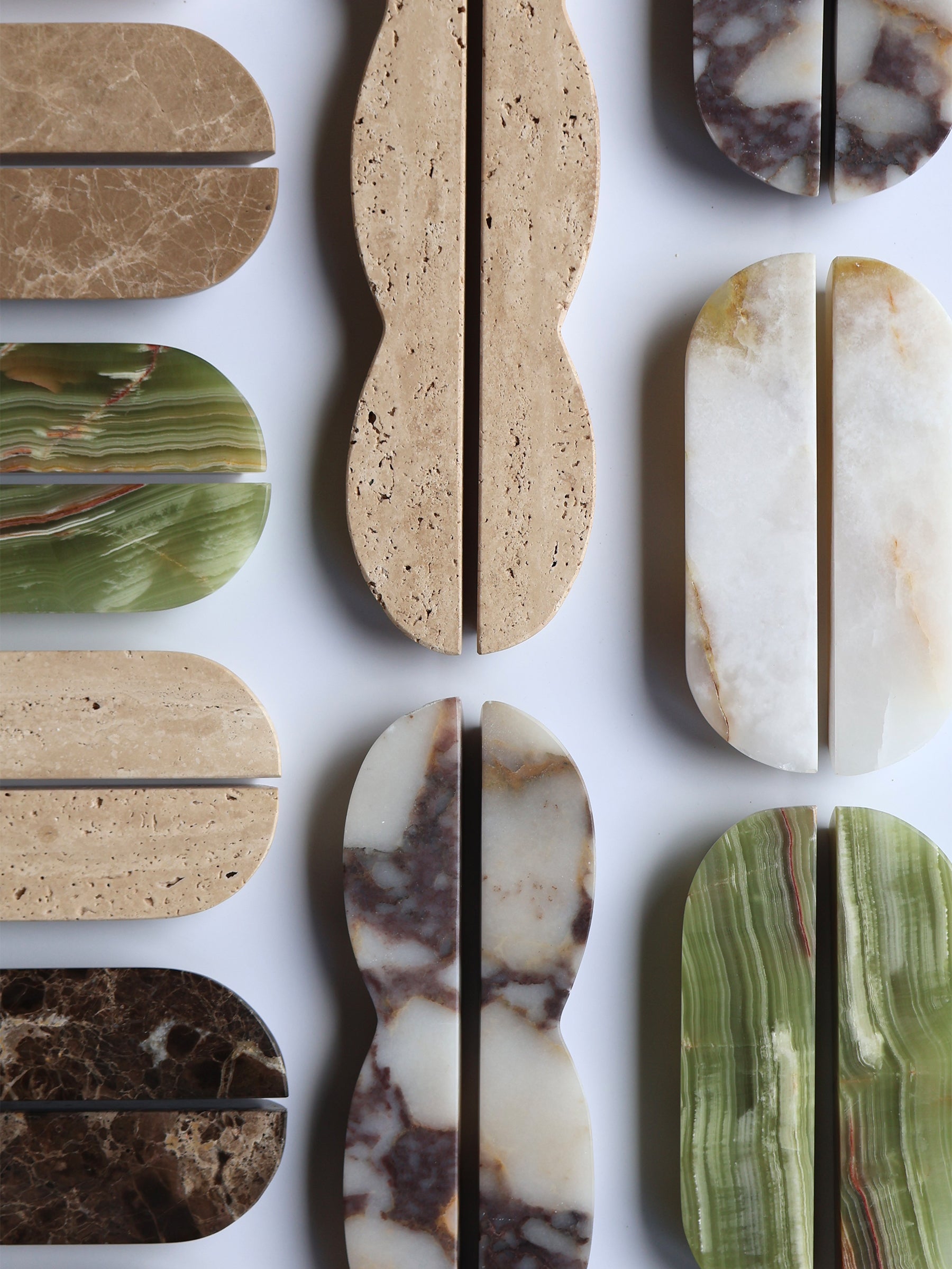 A flat-lay arrangement of polished stone and marble material samples, each cut vertically into two pieces, showing varied colors and textures including beige travertine, green agate, white marble, and dark brown veining on a white background.