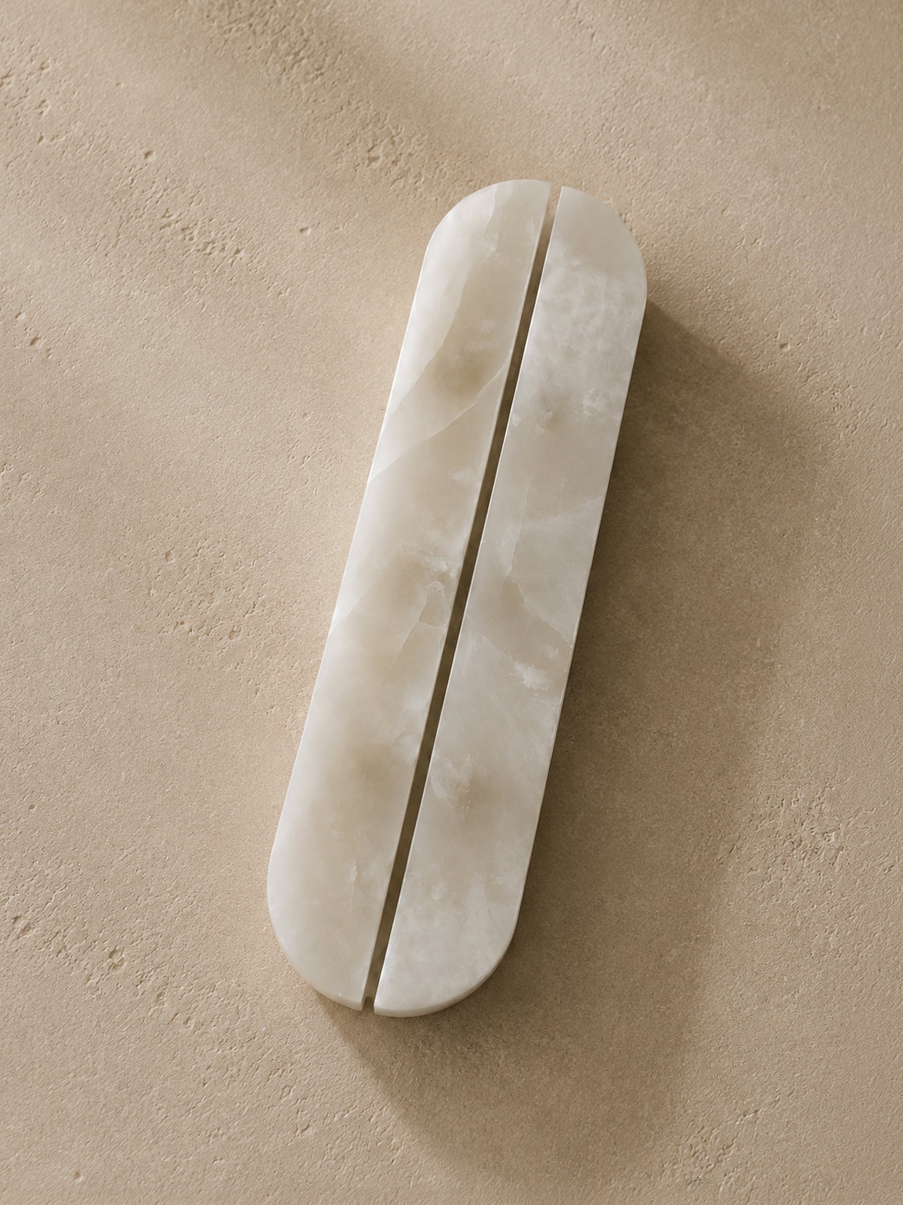 A long, narrow oval bar with a central slit, made of a smooth off-white stone or resin, placed diagonally on a textured beige surface.