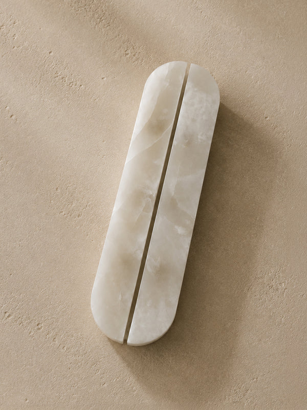 A long, narrow oval bar with a central slit, made of a smooth off-white stone or resin, placed diagonally on a textured beige surface.