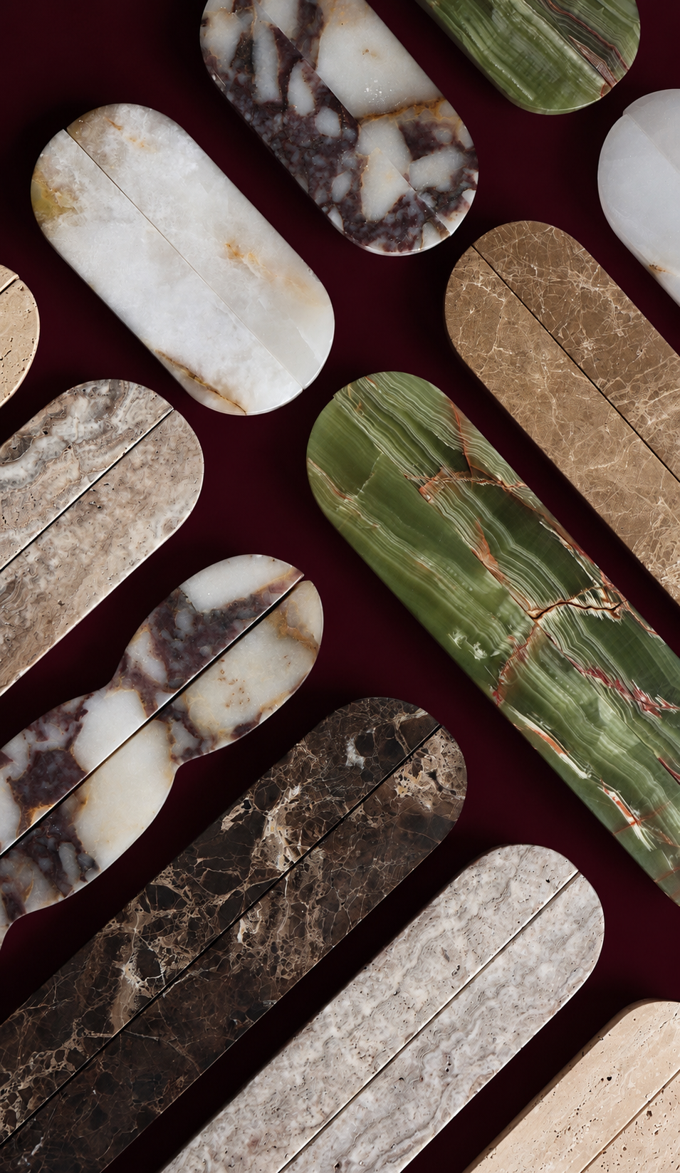 A flat-lay arrangement of elongated oval stone slabs in various colors and marbled patterns, including green, brown, beige, and white, set against a dark burgundy background.