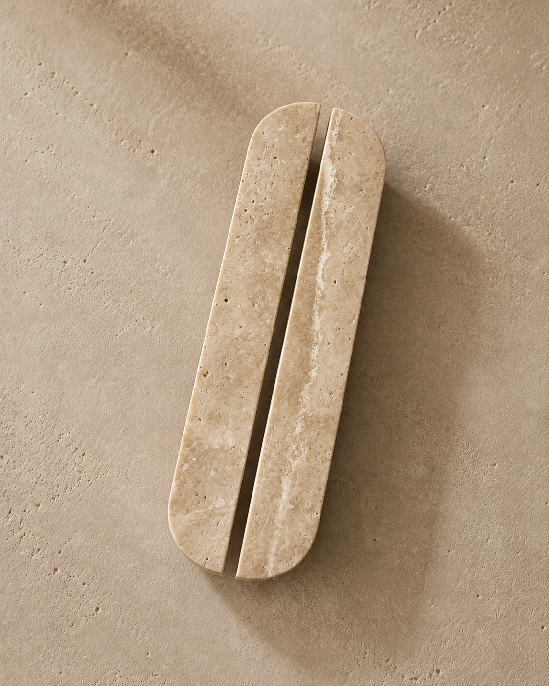 A minimalist wall-mounted soap dish with two elongated rounded rectangular bars separated by a central groove, crafted from light beige travertine stone, shown on a textured beige concrete surface.