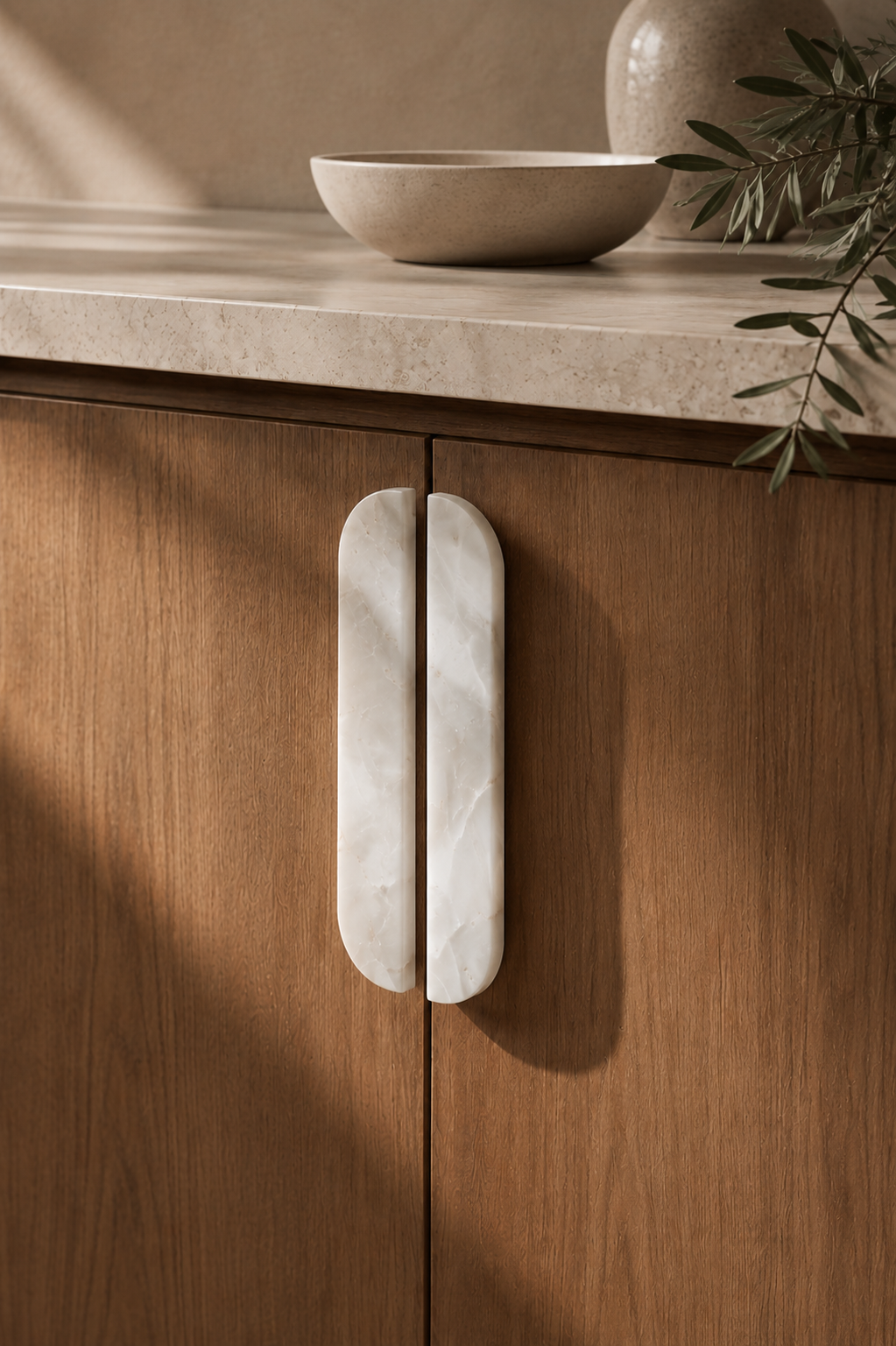 Two elongated white marble cabinet handles mounted on warm brown wooden doors, with a beige stone countertop, a ceramic bowl, and greenery in the background.